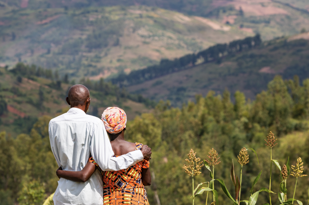 An african couple photographed from behind look out from a hilltop across other hills and scenery. The photograph is in fill colour.